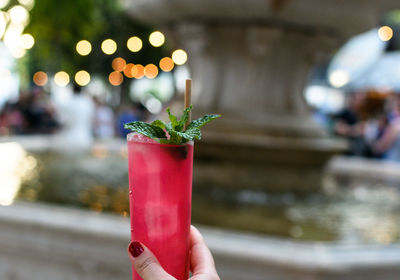 Personal perspective of woman holding red strawberry mojito cocktail in city.