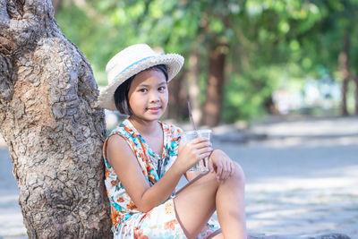 Portrait of girl having drink while sitting by tree trunk