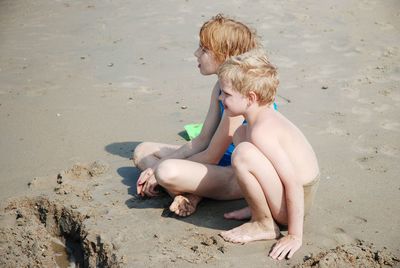 High angle view of siblings looking away while sitting on sand at beach