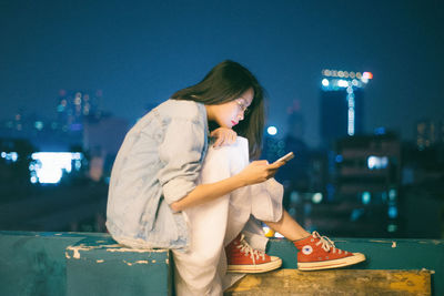 Young woman using mobile phone while sitting on camera at night