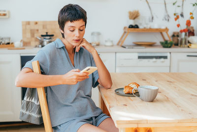 Young european woman sitting in the kitchen and having breakfast uses a mobile phone to communicate