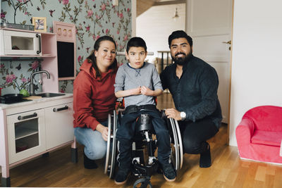 Portrait of smiling parents crouching by autistic son sitting on wheelchair at home