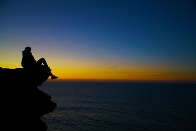 Silhouette woman sitting on rock by sea against sky during sunset