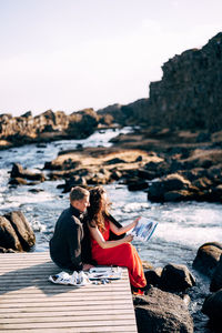 People sitting on rock by sea against sky