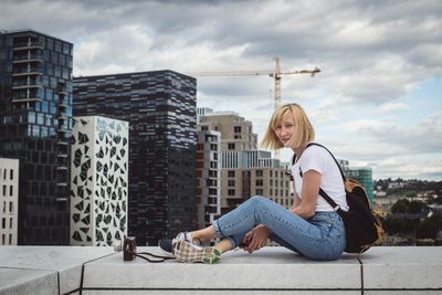 Young woman sitting on retaining wall against cityscape