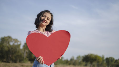 Portrait of a smiling young woman holding heart shape against sky