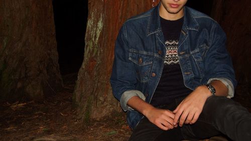 Midsection of young man sitting by tree on field at night