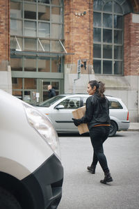 Full length of female messenger carrying box while crossing road amidst cars in city