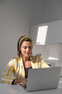 Positive busy pregnant female remote worker in stylish striped jacket and headband sitting at table and browsing laptop while working on project at home