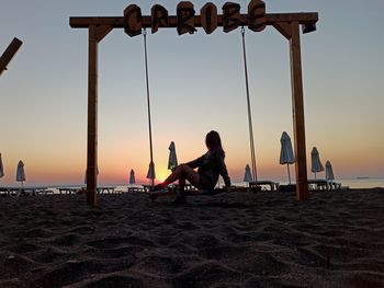 People sitting on swing at beach against sky during sunset
