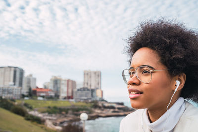 Young woman looking away while listening music against sky during day