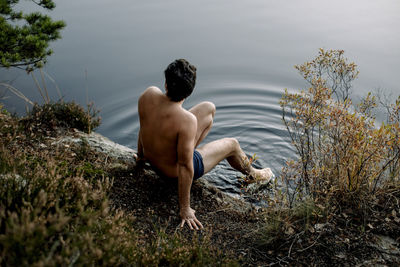 Shirtless man sitting near lake with legs dipped in water