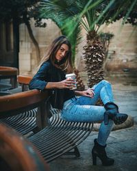 Full length portrait of young woman holding disposable cup while sitting on bench