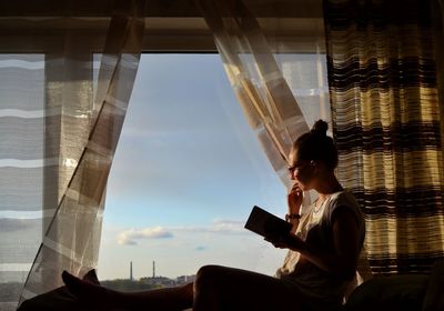 Side view of woman with book sitting on window at home