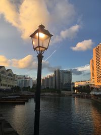 Illuminated street light by river against buildings in city at dusk