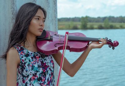 Young woman playing violin by river while standing against river