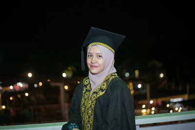 Smiling young woman wearing mortar board while standing against sky at night