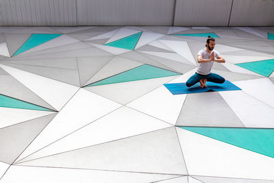 Bearded guy in sportswear practicing squatting on tip toe balance with prayer mudra gesture in spacious room with geometric wall and floor