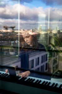 A boy playing  the piano in a window with the reflection of a townscape in it 