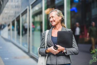 Happy mature businesswoman looking away while standing against office building