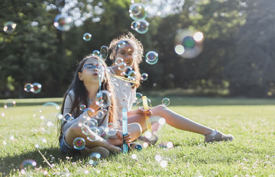 Two caucasian girls sisters blowing out a lot of soap bubbles while sitting in the park