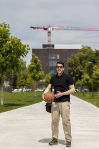 Full length of man holding basketball while standing on footpath against sky