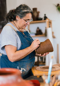 Mature female craftswoman ceramist in apron using tool and creating pattern on clay pot in workshop