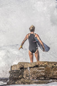 Rear view of woman with surfboard standing on rock at sea