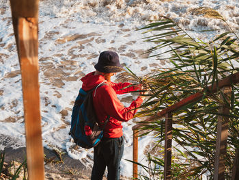 Bearded man in red hoody with backpack standing against sea with reeds. millennial guy travel alone.