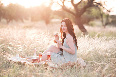 Happy young woman having picnic in meadow with glass of wine, fruit over sunset outdoors. 