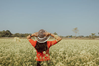 Full length of woman in a hat standing on a yellowish mustard field against clear sky. 