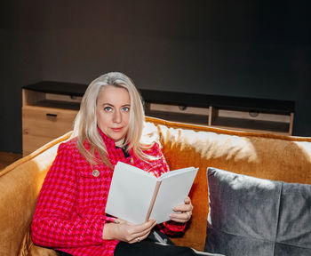 Elegant woman in vibrant red jacket reading a book on sofa