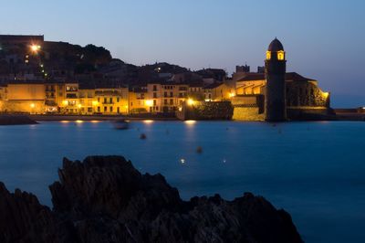 Illuminated buildings by sea against sky at dusk