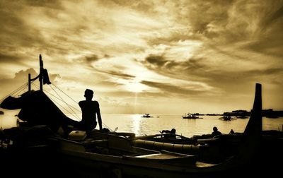 Silhouette man sitting on moored boat at beach against sky during sunset