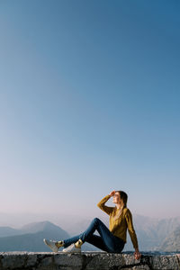 Side view of woman sitting on rock against clear sky