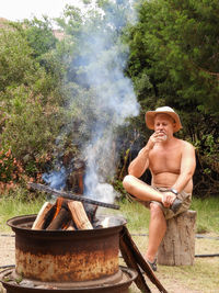 Full length of shirtless man smoking cigarette while sitting by fire pit against trees