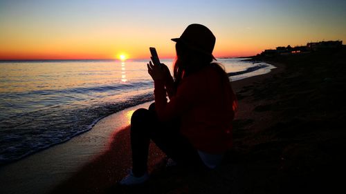 Woman using mobile phone at beach against sky during sunset