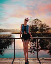 Full length of woman standing by railing against lake during sunset