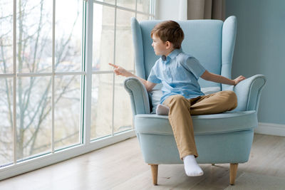 Cute boy sits in chair by window and looks out for something in window