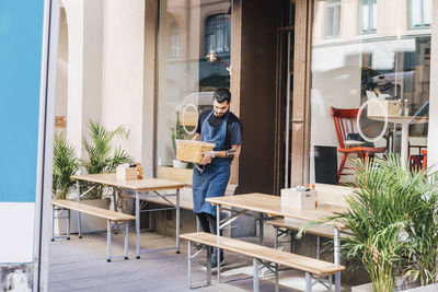 Full length of young male owner carrying food container amidst furniture against restaurant