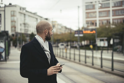 Mid adult businessman holding smart phone while standing on sidewalk in city