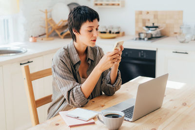 Young caucasian modern female freelancer sitting in the kitchen uses mobile phone and computer.