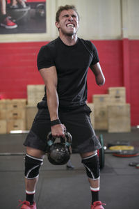 Full length of aggressive male adaptive athlete lifting kettlebell while standing against wall in gym