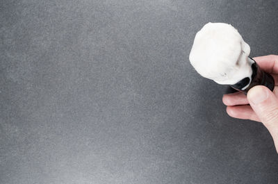 Cropped hand of man holding shaving brush with cream by gray wall