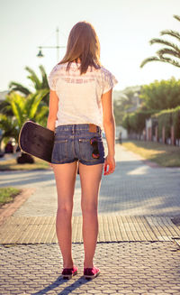 Back view of beautiful young girl with short shorts and skateboard outdoors on a hot summer day.