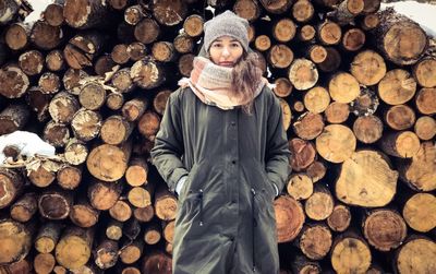 Portrait of woman with hands in pockets standing against logs during winter