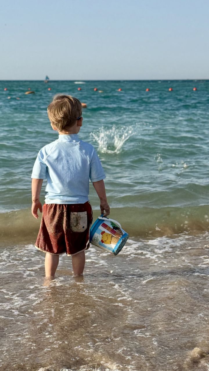 water, sea, beach, vacation, rear view, childhood, child, land, shore, body of water, ocean, one person, full length, nature, horizon over water, sand, horizon, men, coast, sky, wave, leisure activity, day, holiday, trip, casual clothing, standing, beauty in nature, lifestyles, summer, outdoors, sunny, motion, sunlight, blue, toddler, clear sky, fun, scenics - nature, looking, clothing