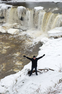 High angle view of man with arms outstretched standing on snow covered field by waterfall