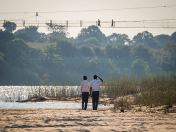Rear view of two people walking on river shore against sky and footbridge, chinyingi, zambia