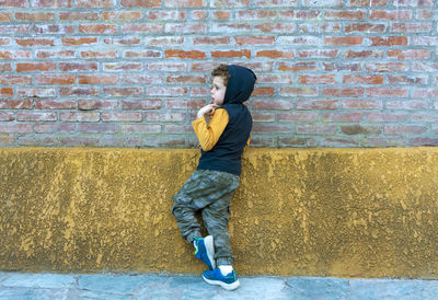 Child in hooded sweatshirt and camouflage pants leaning against brick wall